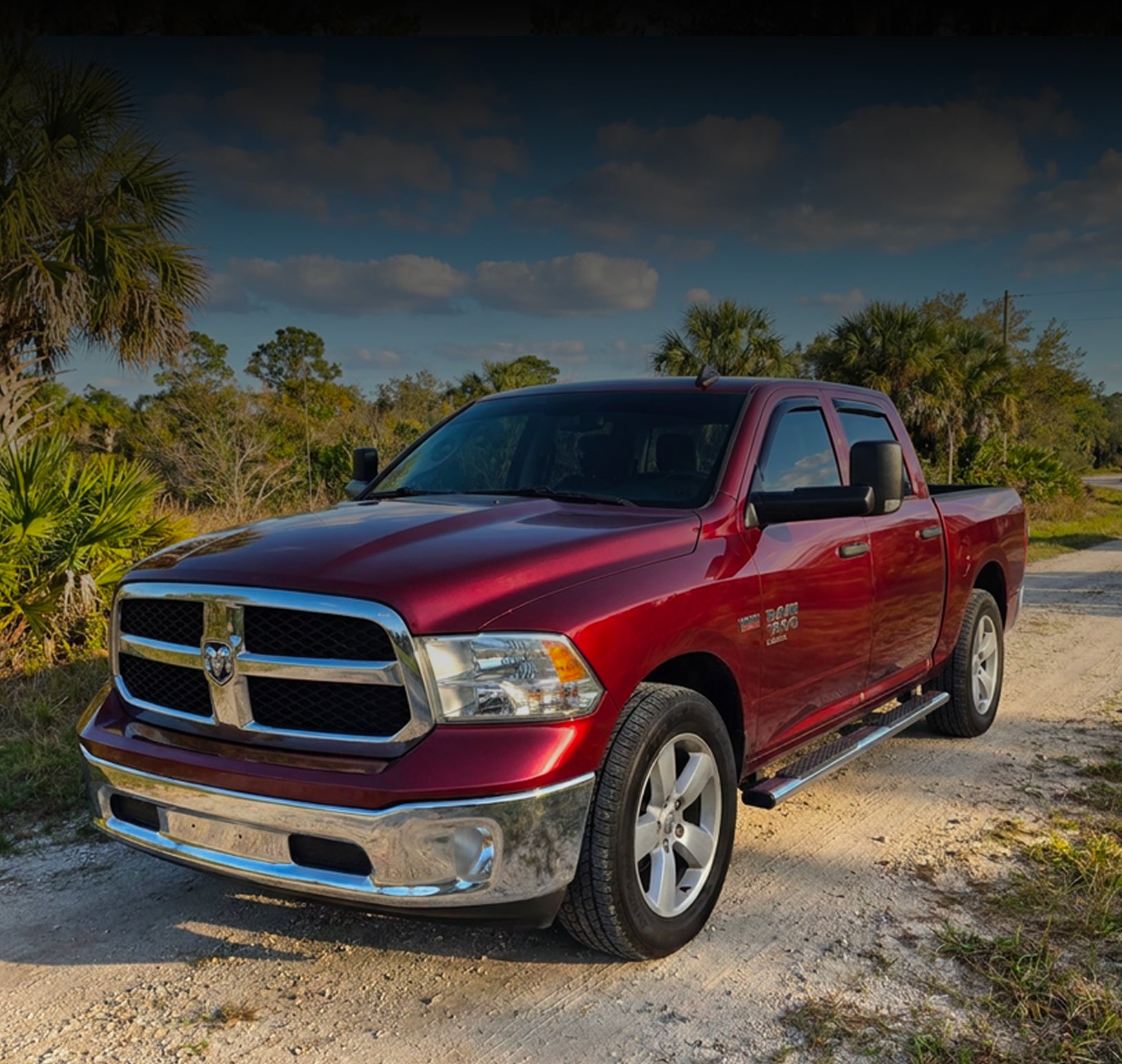A red Ram pickup truck parked on a dirt road outdoors, with trees and a cloudy sky in the background.