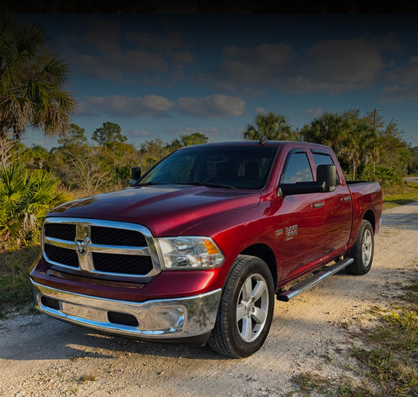 A red Ram pickup truck parked on a dirt road outdoors, with trees and a cloudy sky in the background.