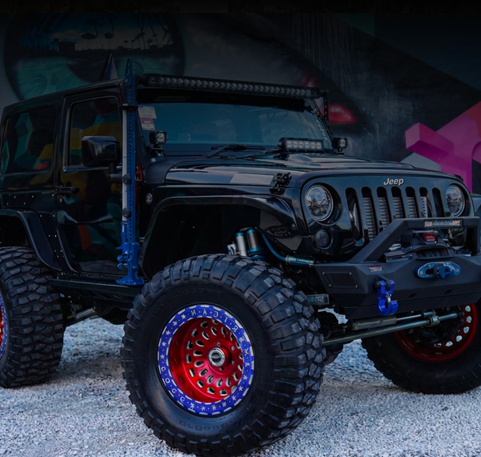A black lifted Jeep Wrangler with large off-road tires, red and blue custom wheels, and a front bumper brush guard, parked on gravel in front of a dark graffiti wall.