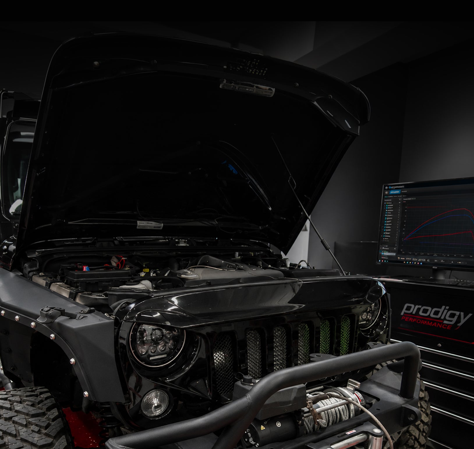 Jeep with open hood in a garage setting, featuring a computer monitor displaying 'prodigy'.